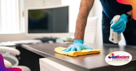 A man wiping a desktop with a yellow cloth holding a spray bottle in the other hand